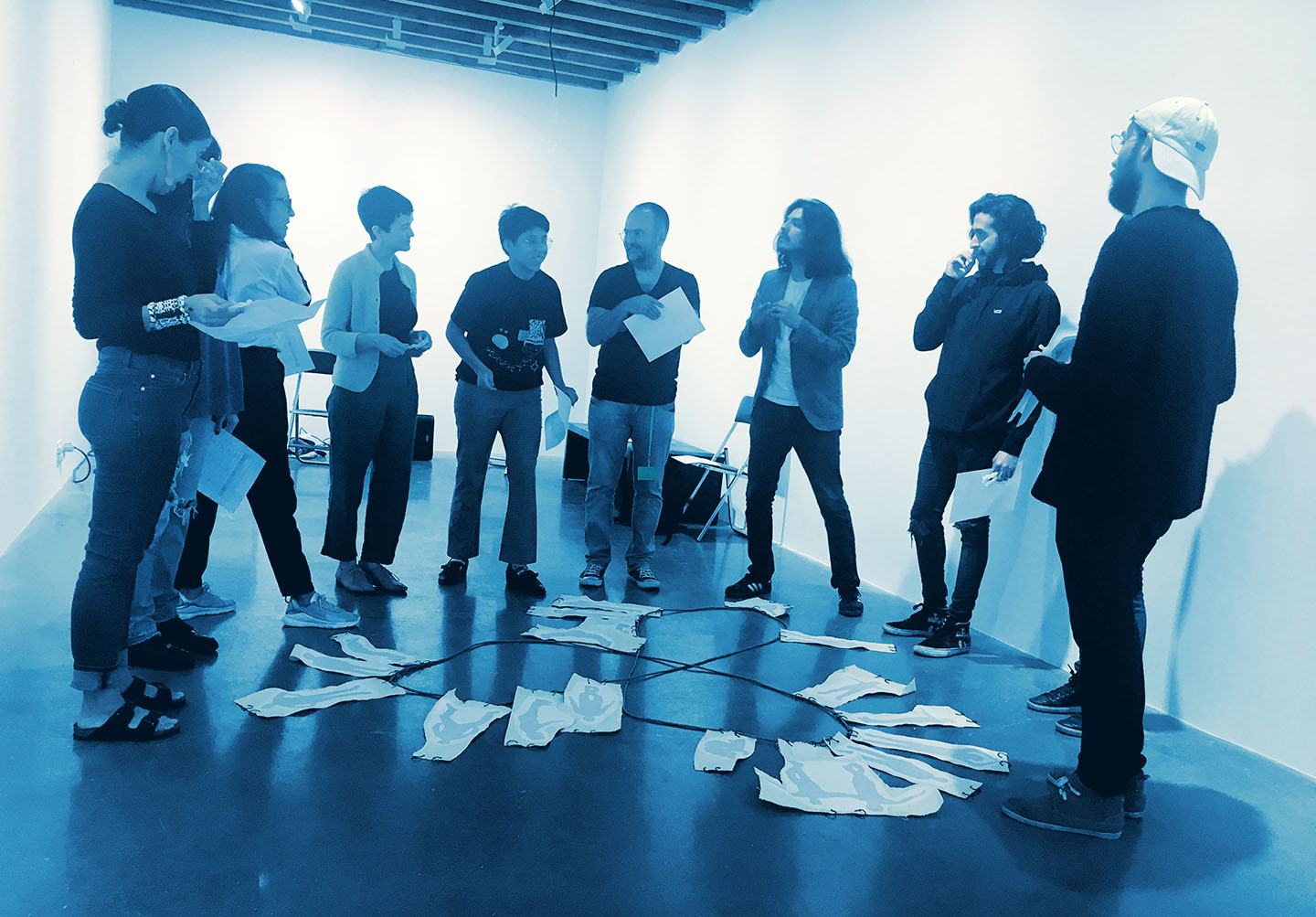Ten people standing and speaking, in a semi-circle around a diagrammatic instrument on the ground. The diagrammatic instrument is a large trefoil knot on the ground that has images of people in various poses attached to it.